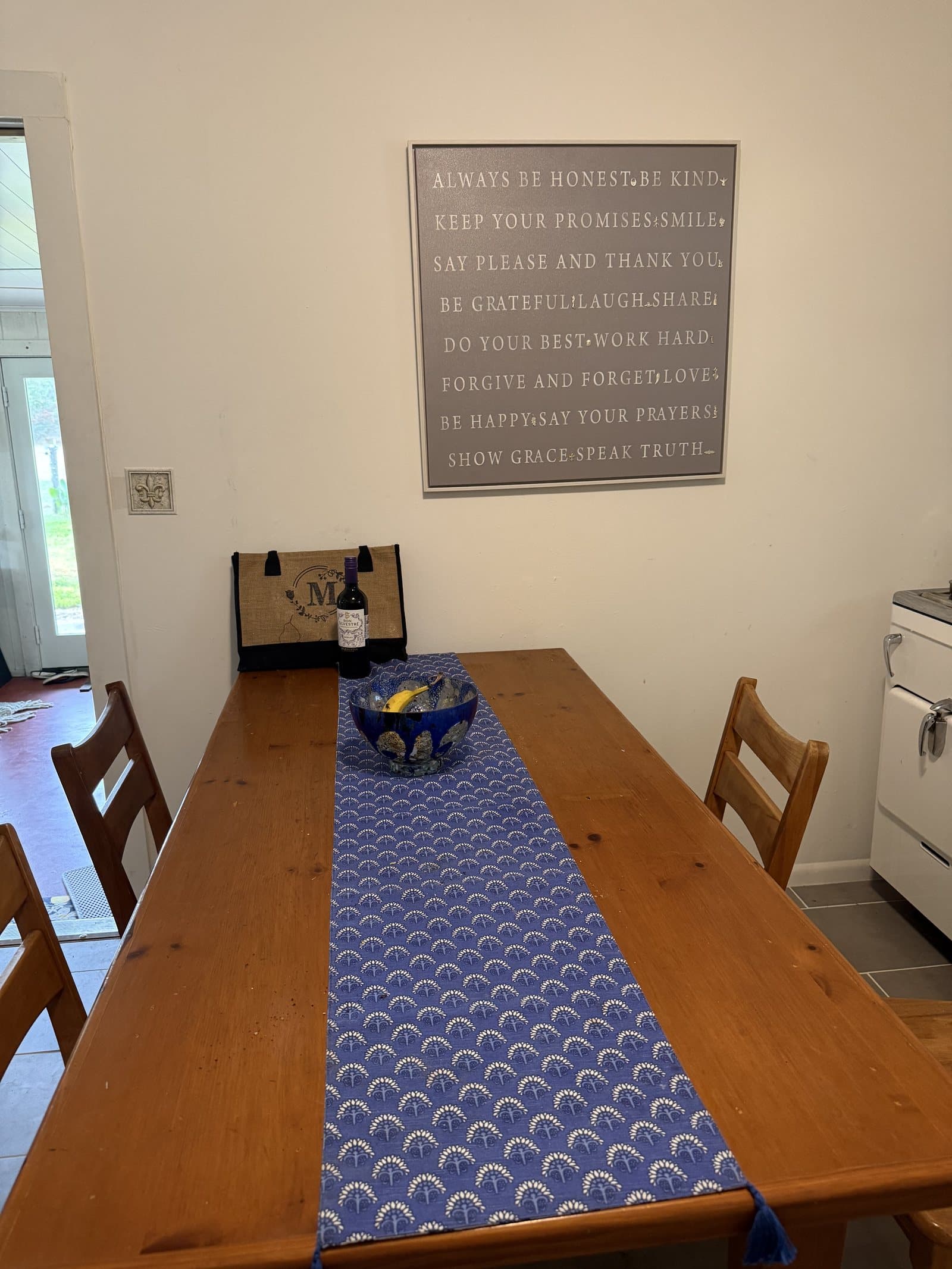A long wooden farmhouse table with a blue runner in the dining area at ROMAR House, beneath a framed sign of house sayings.
