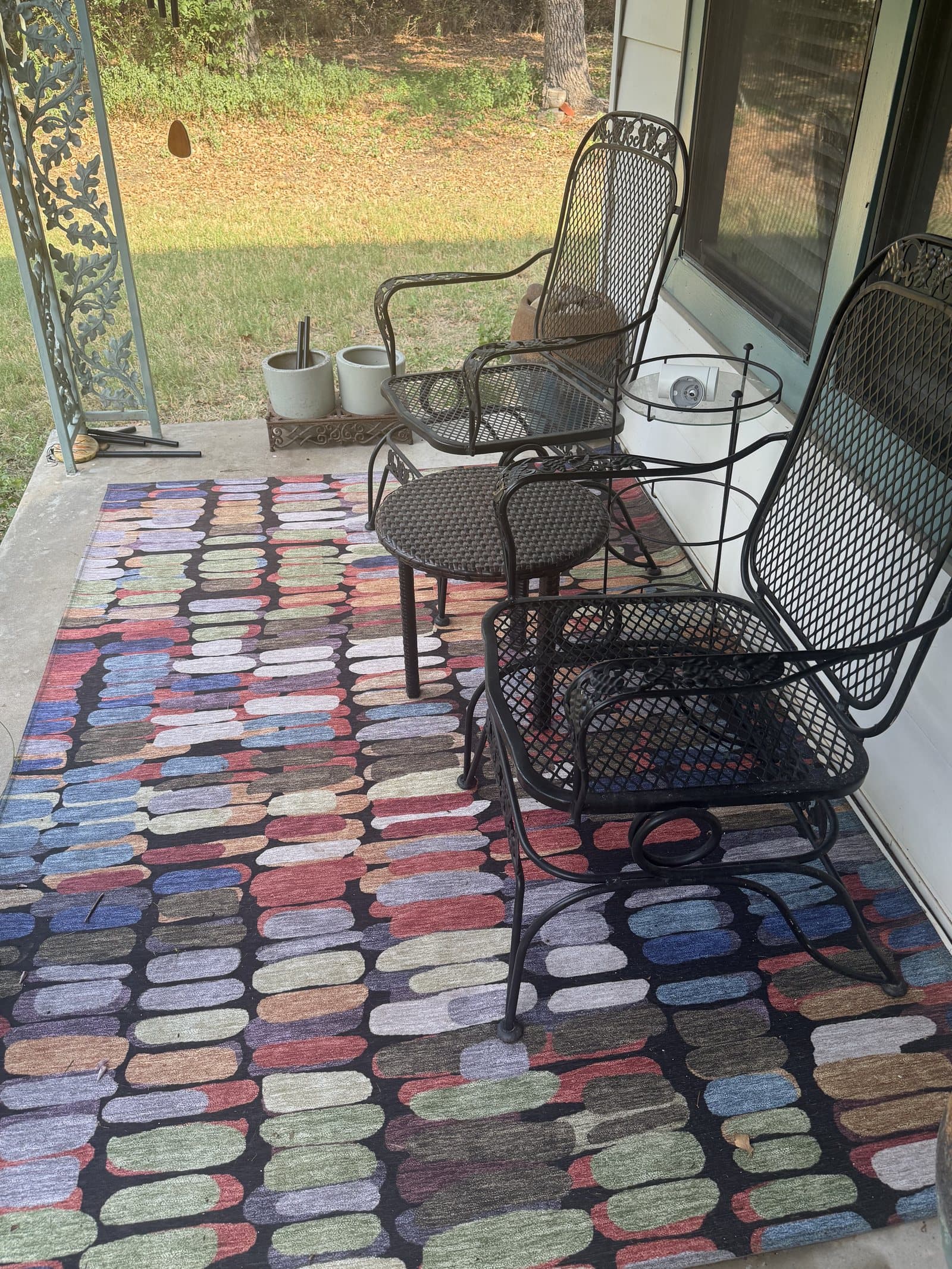 Wrought-iron chairs and a small table on the porch of ROMAR House, on a colourful rug, looking out over the lawn.