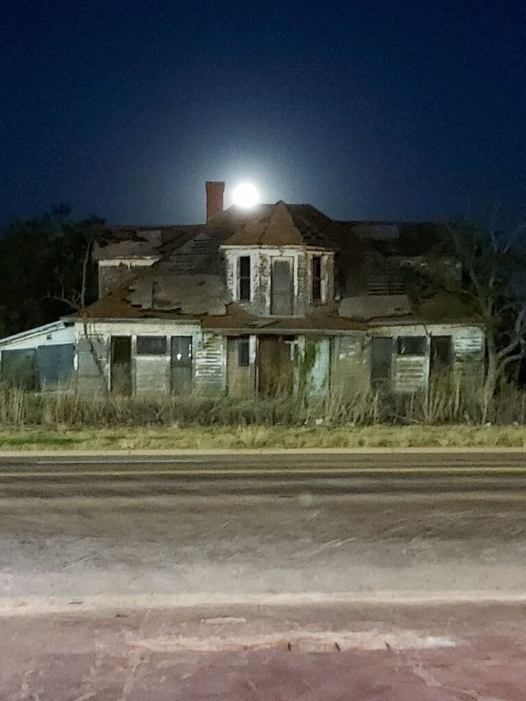 An abandoned two-story house with carved eaves and a sagging roof, under a full moon